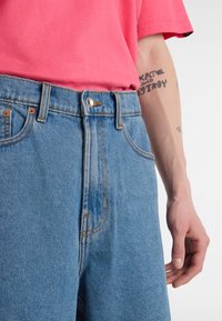 Jasnoniebieskie spodnie dżinsowe z wysokim stanem, ozdobione miedzianymi nitami i detalami szwów. Do tego jasnoróżowa koszula.