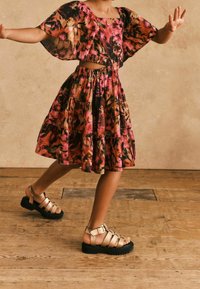 Farbiger, gestufter Kleid in einem floralen Muster aus Pink, Orange und Schwarz mit weiten Ärmeln. Kombiniert mit goldenen Riemchensandalen auf einem Holzboden.