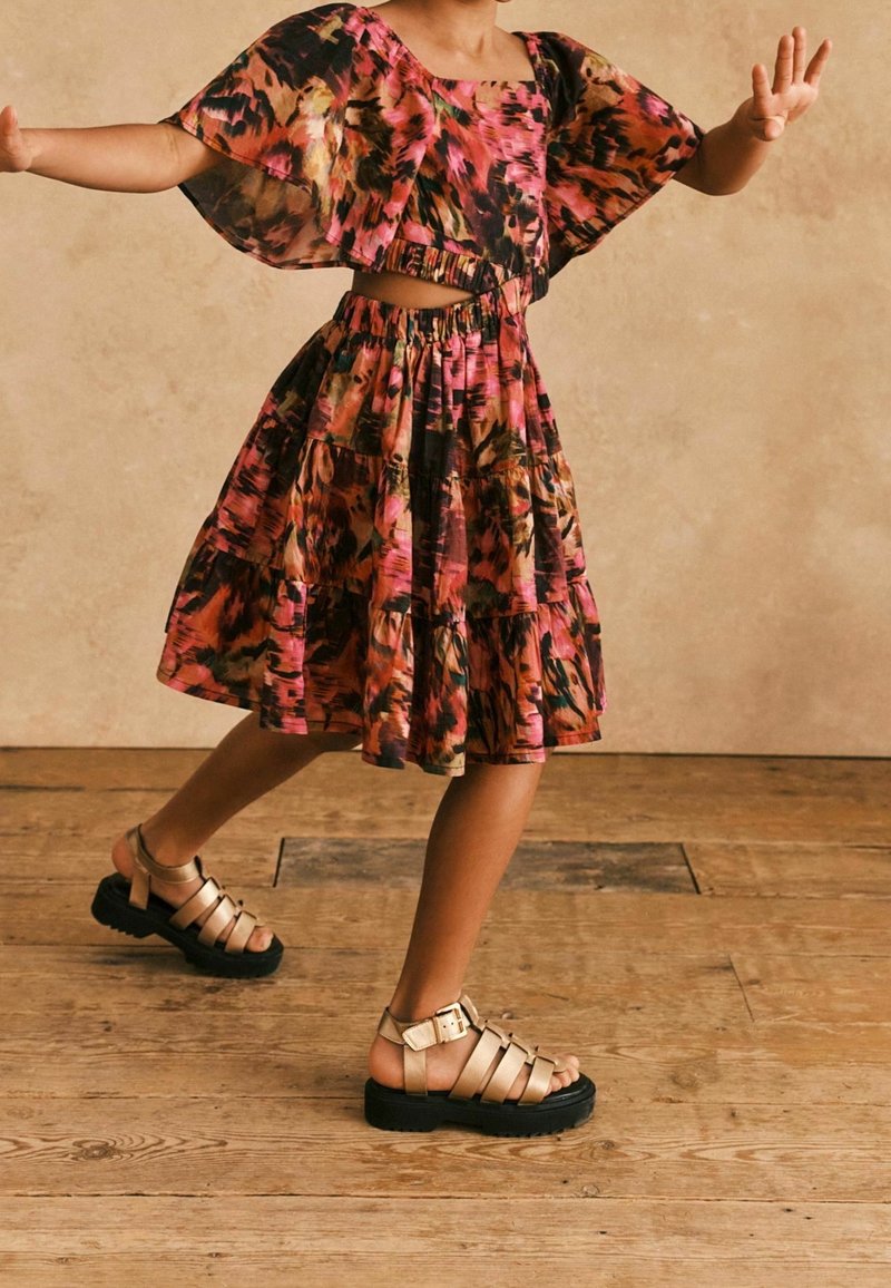 Farbiger, gestufter Kleid in einem floralen Muster aus Pink, Orange und Schwarz mit weiten Ärmeln. Kombiniert mit goldenen Riemchensandalen auf einem Holzboden.