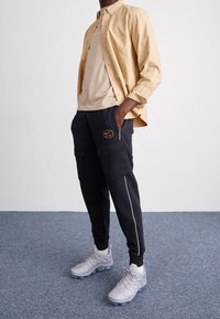 Camisa de botones beis, camiseta de color marrón claro, pantalones jogger de cargo negros con bolsillos laterales y ribetes blancos, zapatillas gris con detalles texturizados.