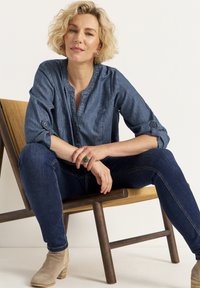 Blaue Denimbluse mit Knopfdetails, kombiniert mit dunklen Jeans und beigen Knöchelstiefeln. Sitzend auf einem Holzstuhl, der einen grünen Ring präsentiert.
