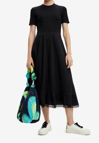 Schwarzes, geripptes Oberteilkleid mit strukturiertem Rock und Spitzenbesatz, kombiniert mit einer bunten, gefalteten Tasche und weißen Sneakern.