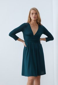 Turkusowa sukienka o kroju kopertowym z rękawami do trzy czwartej, z głębokim dekoltem w kształcie V i zebranym w pasie, wykonana z miękkiego materiału jersey.
