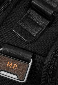 Detalle de mochila de tela negra con hebilla de cuero texturizado con un acento marrón y una placa metálica con las inscripciones "M.P." y "Piquadro".
