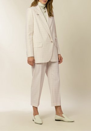 Vrouw draagt crème kleurig geruit pak met blazer, cropped wijde broek, witte coltrui en bijpassende loafers, staand.
