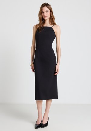 Donna che indossa un abito midi nero aderente con spalline sottili e décolleté nere con punta appuntita, in piedi davanti a uno sfondo grigio chiaro uniforme.