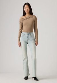 Γυναίκα που στέκεται, φοράει μπεζ τοπ με ανοιχτούς ώμους, ανοιχτό μπλε ψηλόμεσα τζιν και μαύρα μποτάκια με τακούνι σε απλό φόντο.