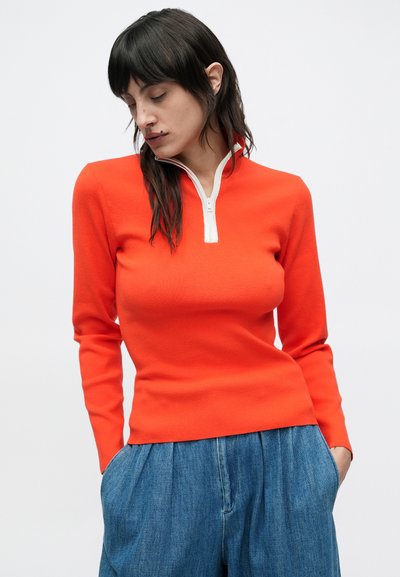 Haut rouge à manches longues avec fermeture éclair blanche, coupe ajustée, texture côtelée. Associé à un pantalon en denim bleu clair, ample. Fond uni.