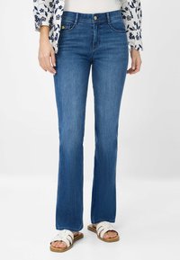 Uiteengelopen blauwe spijkerbroek met een gemiddelde wassing, voorzien van voorzakken, riemlussen en een gladde textuur. Gecombineerd met witte sandalen.