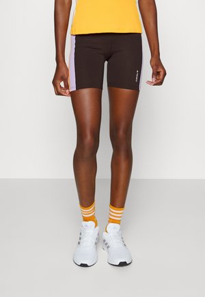 Brune atletiske shorts med lavendel sidepaneler, parret med en gul top og hvide sko. Modellen har også orange stribede sokker på.
