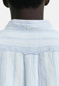 Camisa de rayas azul claro con una textura suave. Presenta un cuello abotonado y rayas verticales con un leve detalle de contraste a lo largo de la espalda.