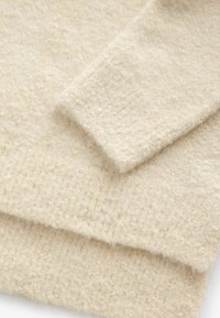 Pull crème réalisé en tissu doux et texturé, avec une surface légèrement duveteuse et un motif côtelé le long de l'ourlet et des manches.