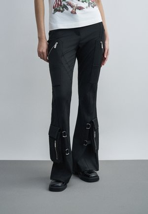 Czarne spodnie typu cargo z wieloma kieszeniami na zamek, klamrami w okolicy kostek, noszone z czarnymi butami i białym topem z nadrukiem ptaka.