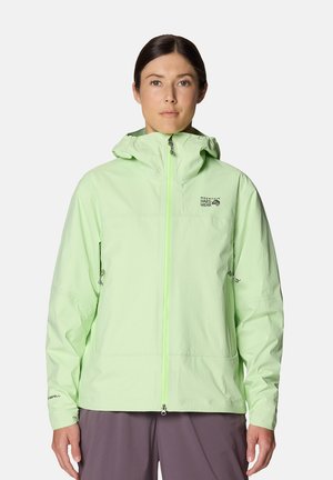 Femme portant une veste d'extérieur à capuche vert clair avec une fermeture éclair devant et un pantalon gris, debout devant un fond uni.