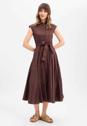 Femme portant une robe midi marron sans manches boutonnée avec une ceinture nouée à la taille et des sandales beiges à bouts ouverts, debout devant un fond blanc uni.