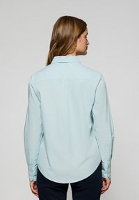 Jasnoniebieska koszula z długimi rękawami, ściąganymi mankietami, ze spiczastym kołnierzykiem oraz detalem w formie yoku na plecach. Gładka tekstura materiału.