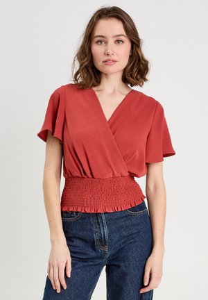 Top avvolgente color ruggine con maniche corte e scollo a V; presenta una vita arricciata con finitura testurizzata. Indossato con jeans blu scuro.