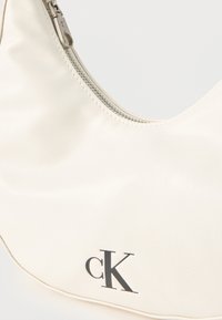 Λευκή δερμάτινη τσάντα με ασημί φερμουάρ και μαύρο λογότυπο CK στο μπροστινό μέρος, κυρτό σχήμα και λεία υφή.