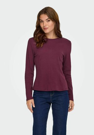 Langarm, getailleerd shirt in donker bordeaux, ronde halslijn, zachte stof met naaddetails; gedragen met donkere jeans.