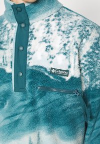 Pull en polaire à motifs bleu sarcelle et blanc avec boutons-pression, poche zippée et logo Columbia Sportswear Company sur la poitrine.