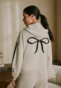 Beige sweater med hætte, med et stort sort sløjfedesign på ryggen og kontrasterende sorte striber på ærmerne. Blød tekstur.