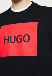 Černá mikina s velkým červeným obdélníkovým potiskem s nápisem "HUGO" tučným černým písmem a žebrovaným výstřihem.