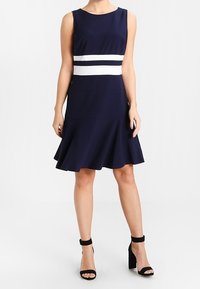 Námořnická modrá bezramenná šaty s bílými horizontálními pruhy v pase a rozšířenou sukní. Měkký materiál a délka po kolena.