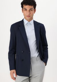 Navy double-breasted blazer with black buttons, structured design, worn over a light blue striped shirt, paired with light gray patterned trousers.
