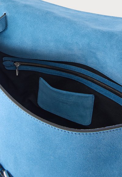 Sac à main en daim bleu ouvert montrant l'intérieur en tissu noir avec poche zippée et une étiquette de marque en daim bleu assortie à l'intérieur.