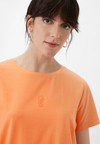 Oranges Baumwoll-T-Shirt mit Rundhalsausschnitt und kurzen Ärmeln, das ein dezentes, geprägtes Logo auf der Brust trägt. Weicher Stoff.