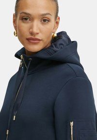 Felpa blu navy con zip frontale, coulisse e cappuccio. Presenta una tasca con zip sulla manica con dettagli in metallo dorato.