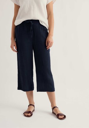 Inimene kannab valget lühikeste varrukatega tekstuurset pusa, tumesiniseid laiade pükstega lühikesi pükse nöörkinnitusega ja pruune lameda tallaga sandaale heledal taustal.