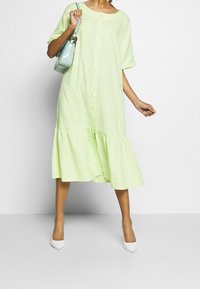 Donna che indossa un vestito midi verde chiaro con bottoni e orlo arricciato, tacchi bianchi a punta e che porta una borsa a tracolla azzurro chiaro.