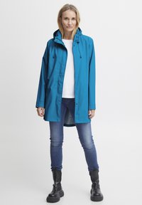 Waterdichte teal regenjas met capuchon, voorkant met drukknoopsluiting, twee zakken en een ontspannen pasvorm, gecombineerd met blauwe jeans en zwarte laarzen.