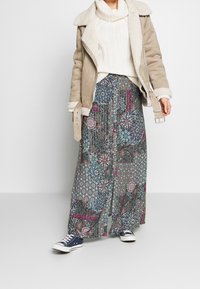 Beige shearling-voering jasje over een witte gebreide trui, gecombineerd met een lange patroonrok in blauwe en roze tinten, en blauwe sneakers.