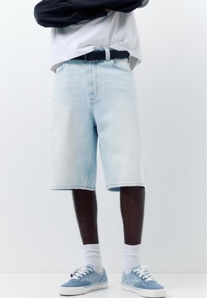 Mężczyzna w jasnoniebieskich dżinsowych krótkich spodenkach, białych skarpetkach, jasnoniebieskich tenisówkach i czarnym pasku, stojący z założonymi rękami.