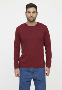 Lange mouwen, bordeauxrood katoenen t-shirt met een ronde halslijn en een klein logo op de borst, gecombineerd met een blauwe spijkerbroek. Gladde textuur.