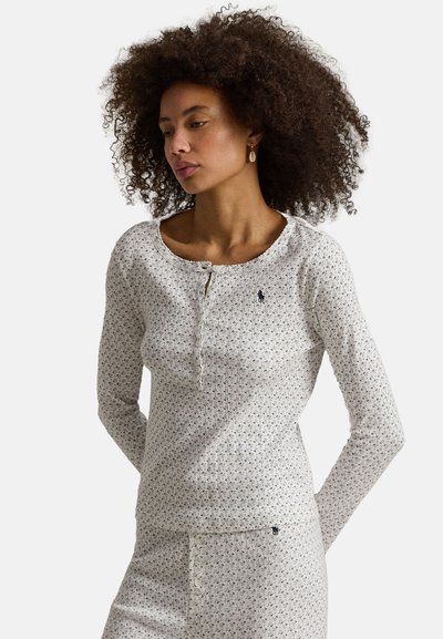 Vrouw met krullend haar, gekleed in een wittig top met lange mouwen met patroon en bijpassende broek, kijkt naar beneden en opzij met de handen achter de rug.