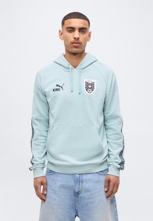 Jeune homme debout portant un sweat à capuche bleu clair avec les logos Puma et ÖFB, associé à un jean bleu clair, sur un fond blanc uni.