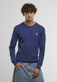 Navy blue sweatshirt in soft fabric, featuring long sleeves and a crew neck. Small white anchor logo on the chest. Dark blue denim skirt.