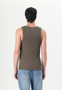 Barna bordázott ujjatlan felső, lekerekített nyakkivágással, slim fit szabással és logó részlettel a hátsó tetején, világoskék farmernadrággal párosítva.