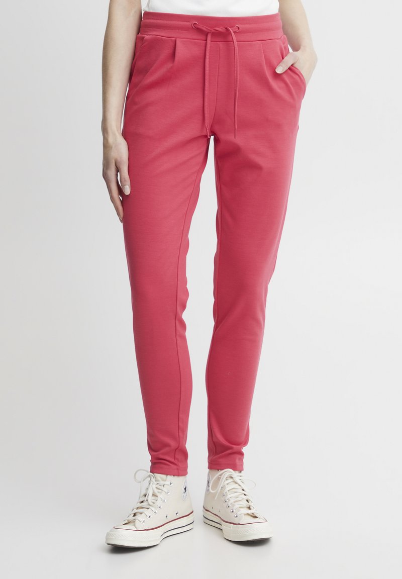 Pink sweatpants with a drawstring waistband, side pockets, and tapered legs, paired with white high-top sneakers. Smooth fabric texture.