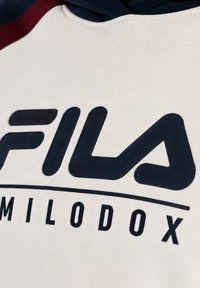 Gros plan d’un vêtement blanc avec un grand logo FILA bleu marine et un texte MILODOX plus petit sous une ligne horizontale.