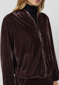 Sieviete valkā tumši brūnu samta džemperi ar rāvējslēdzēju, priekšējām kabatām un elastīgu apakšu, virs melnas augstos kakla džempera.