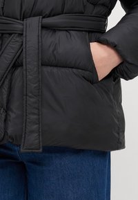 Čierna prešívaná bunda s viazaným pásom, ktorý sa zaviaže v páse, so štruktúrovanou textúrou a bočnými vreckami. Obraz sa zameriava na predný detail bundy a materiál.