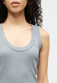 Ženska s kodrastimi lasmi, oblečena v svetlo modro rebrasto majico brez rokavov, vidna od ramen do sredine prsi, na enobarvnem belem ozadju.