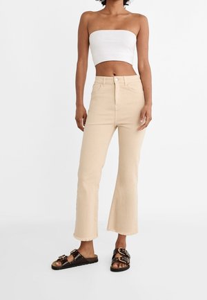 Femme portant un crop top blanc sans bretelles, un jean flare beige raccourci avec des ourlets bruts, et des sandales noires à double boucle, debout sur un fond blanc.
