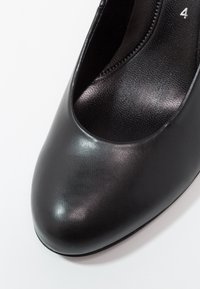 Chaussure à talon haut noire avec une extérieur en cuir synthétique lisse, bout arrondi, et un empeigne basse dévoilant le pied. Doublure intérieure texturée.