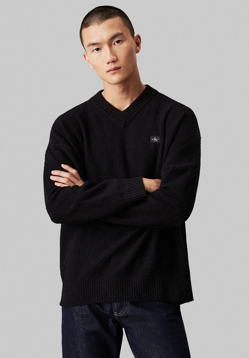 Pull noir à col en V en tissu tricoté texturé avec des poignets et un ourlet côtelés. Présente un petit patch logo sur la poitrine. Porté avec un jean foncé.