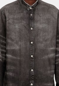 Chemise en denim noir avec col mandarin, fermeture à boutons sur le devant et finition texturée. Présente des boutons argentés et des bords effilochés.